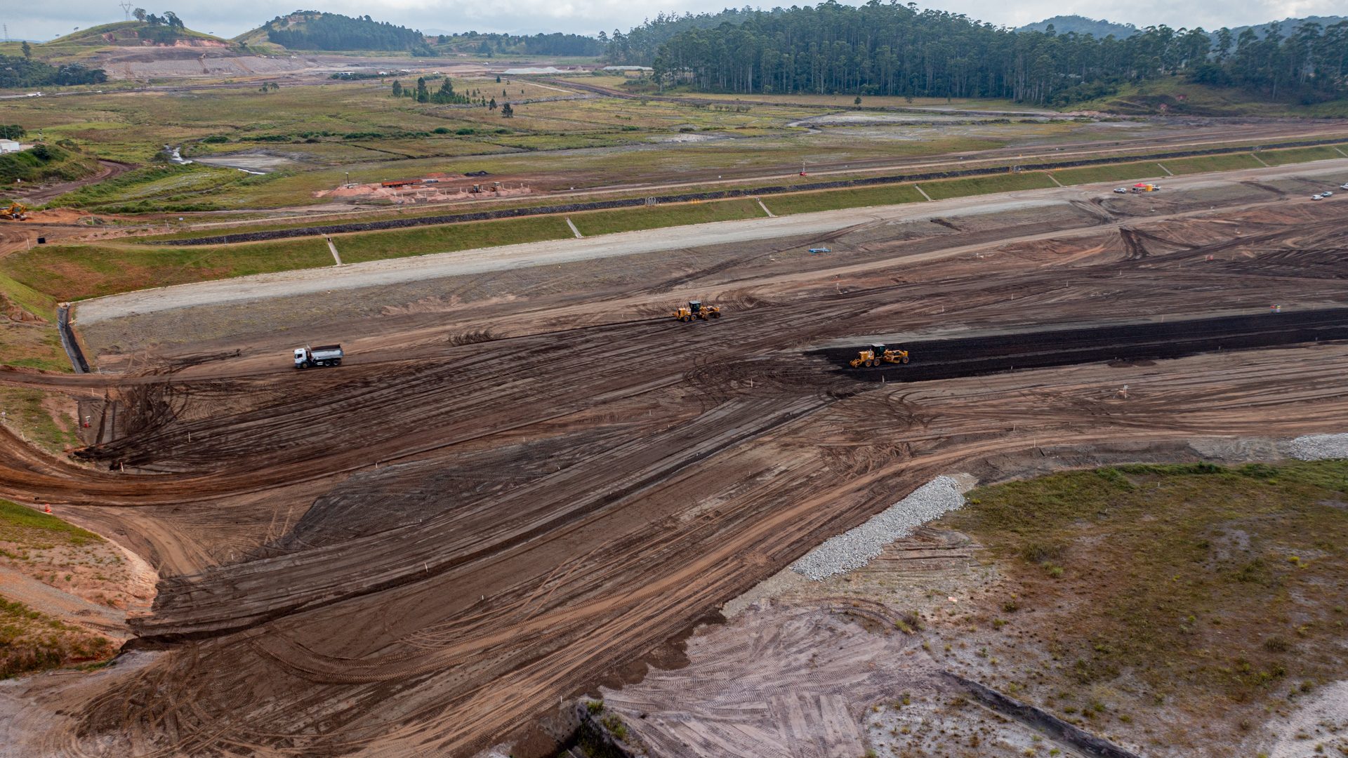 Imagem aérea do Dique 2 em obras. Ao fundo há uma área de vegetação e em primeiro plano uma área terrosa com caminhões trabalhando.
