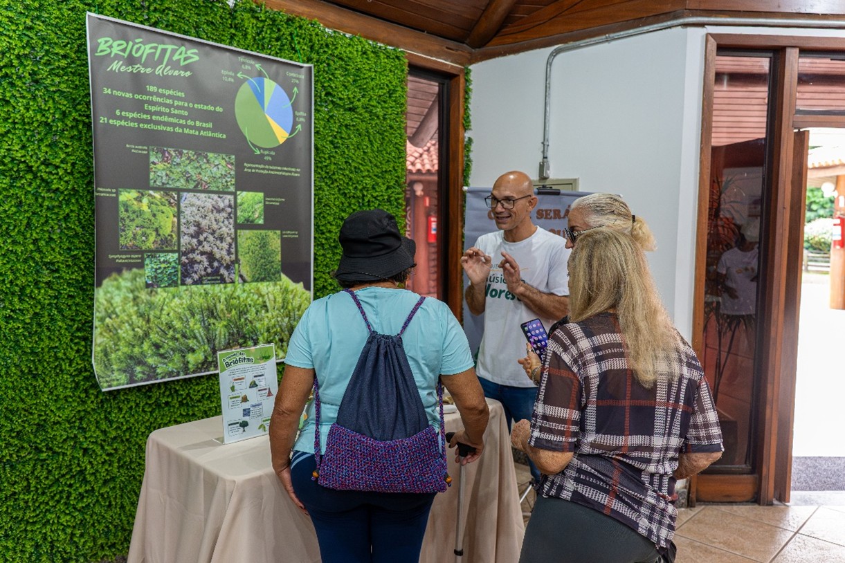 Three women look at a man explaining an educational display about bryophytes at a booth, with a green decorative wall in the background, indoors.