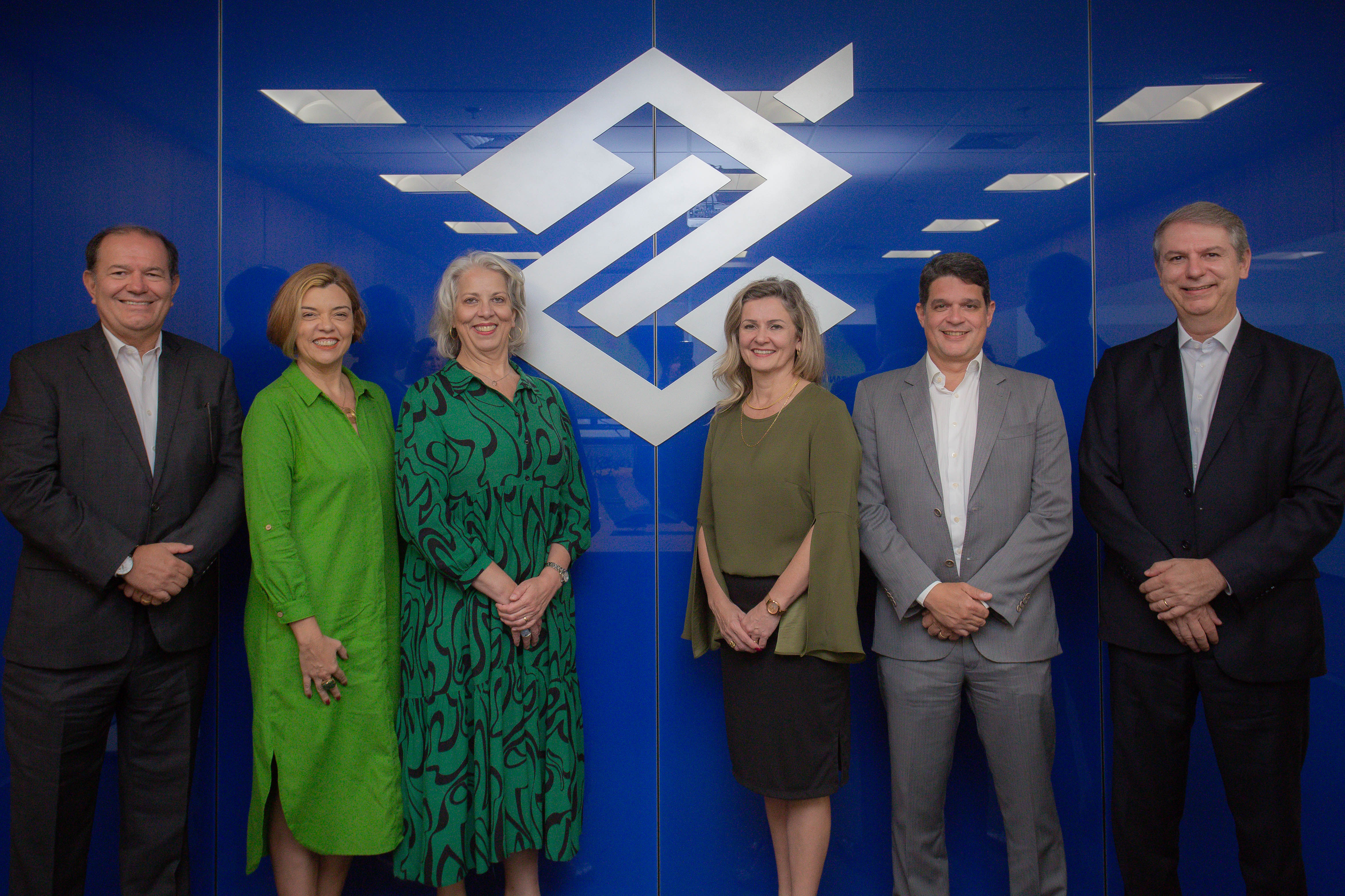 Foto de 3 homens e 3 mulheres na frente de uma parede com símbolo do Banco do Brasil. Da esquerda para direita, tem um homem de blazer, calça social grafite e camisa branca, ao lado uma mulher de vestido longo verde de botões e cabelo loiro curto, ao lado, uma mulher de cabelo branco e vestido verde com linhas azuis. Da direita para esquerda, um homem grisalho de blazer, calça social preta e camisa branca, ao lado um homem de cabelo curto escuro de blazer, calça social cinza e camisa branca, ao lado uma mulher loira com cabelo na altura do ombro, de saia preta e camisa verde de manga longa. 