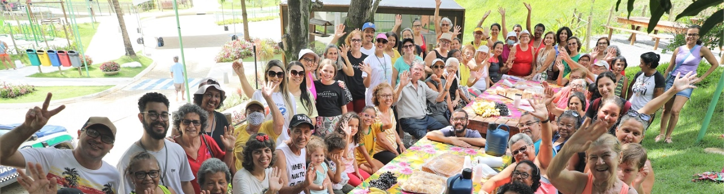 Foto de várias pessoas de diferentes idades ao redor de uma mesa grande retangular, algumas pessoas estão em pé e outras sentadas em bancos, olhando e sorrindo para foto.