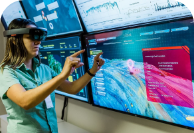 A woman standing in front of some big TVs, wearing virtual reality goggles. They are wearing the Vale uniform and badge.