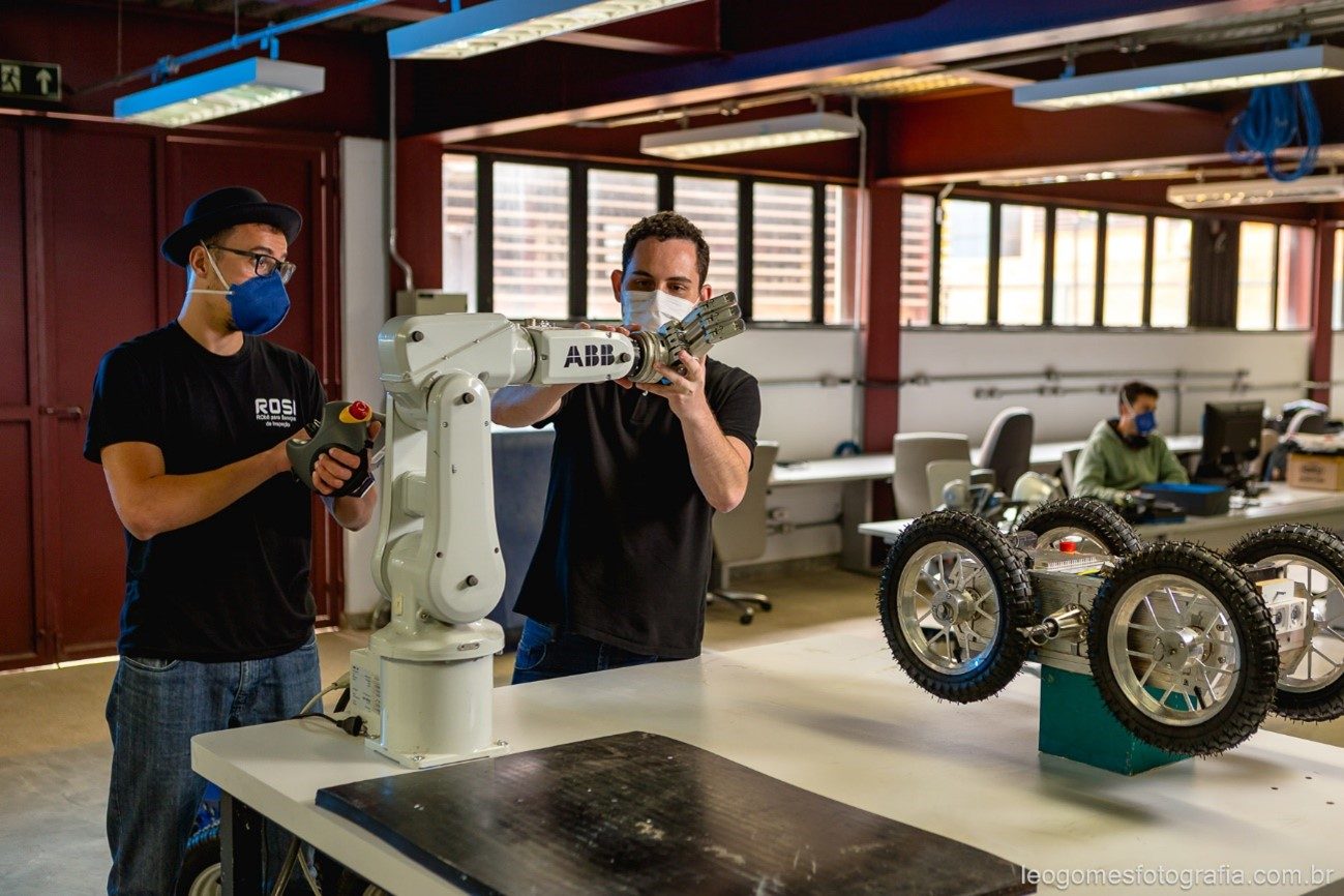 foto de dois jovens mexendo em uma máquina, que está em cima de uma mesa  junto de outros objetos. Ao fundo um outro jovem sentado em uma mesa mexendo no computador. Os jovens em pé estão usando calça jeans, camisa de malha e máscara no rosto.