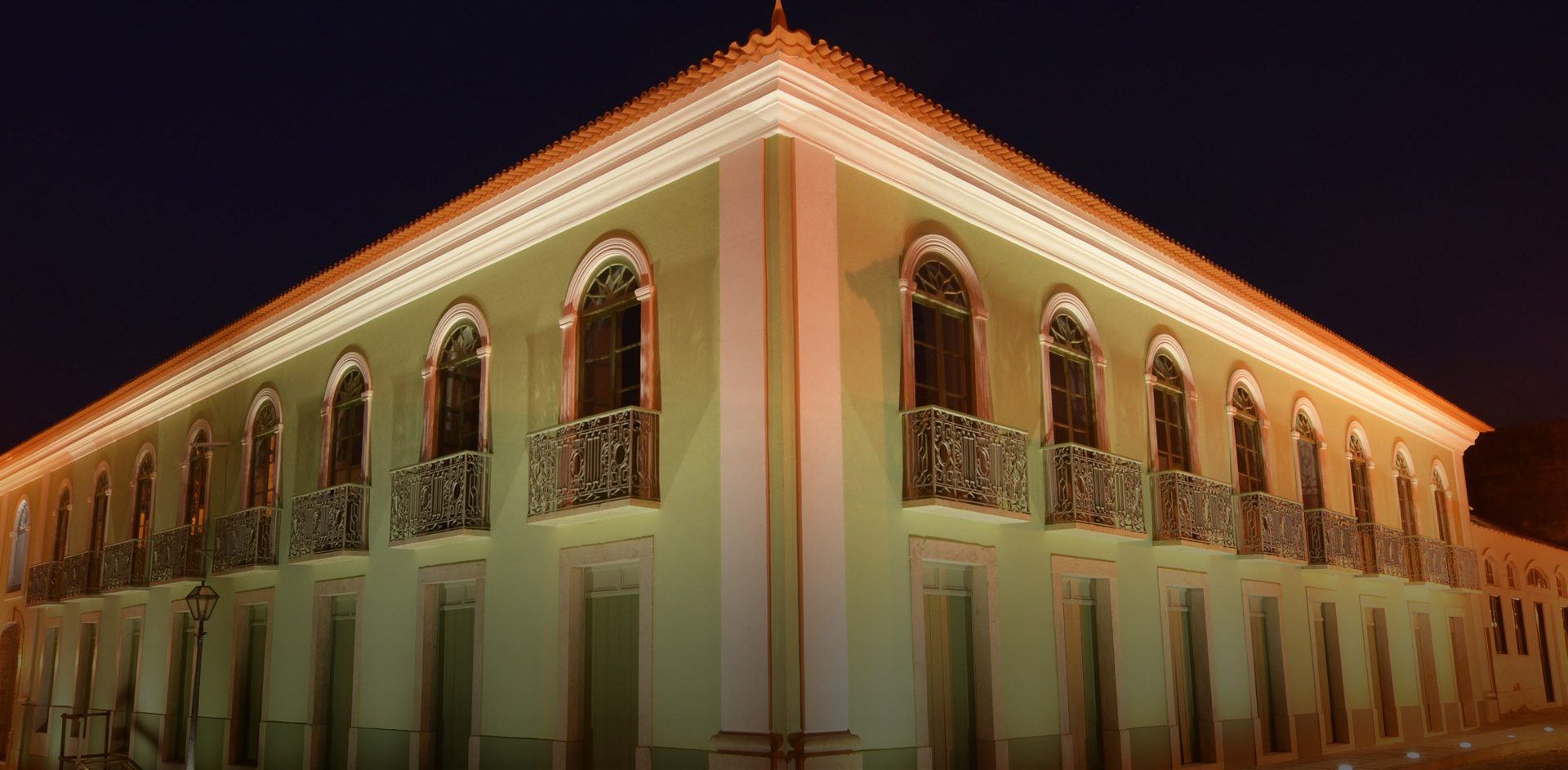 Foto da fachada do Centro Cultural Vale do Maranhão à noite. A foto pega o ângulo de uma quina e dois lados da fachada, que é verde, tem dois andares. O andar de baixo tem várias portas e o andar de cima tem várias janelas com pequenas sacadas.