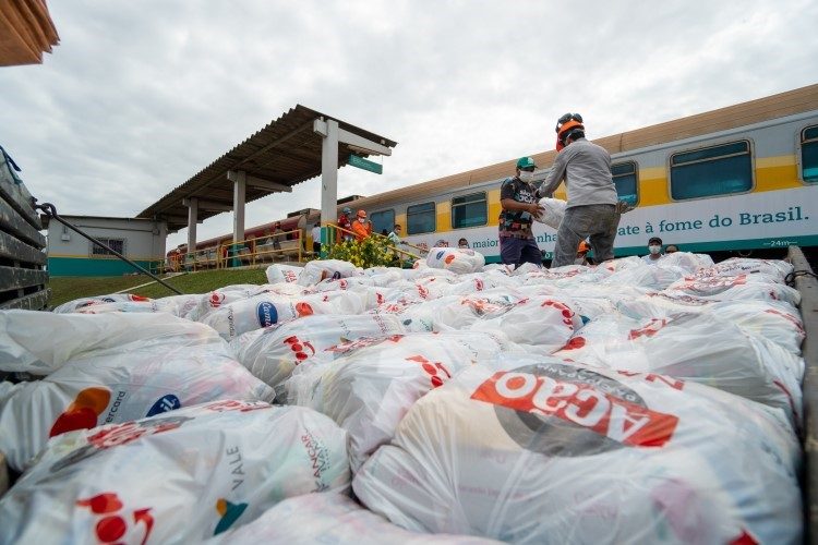 Dois homens entre várias cestas básicas, entregando para algumas pessoas. Ao fundo uma estação de trem e um trem passando.