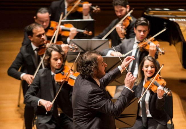 Um maestro rege a orquestra. Há diversos músicos tocando violino 