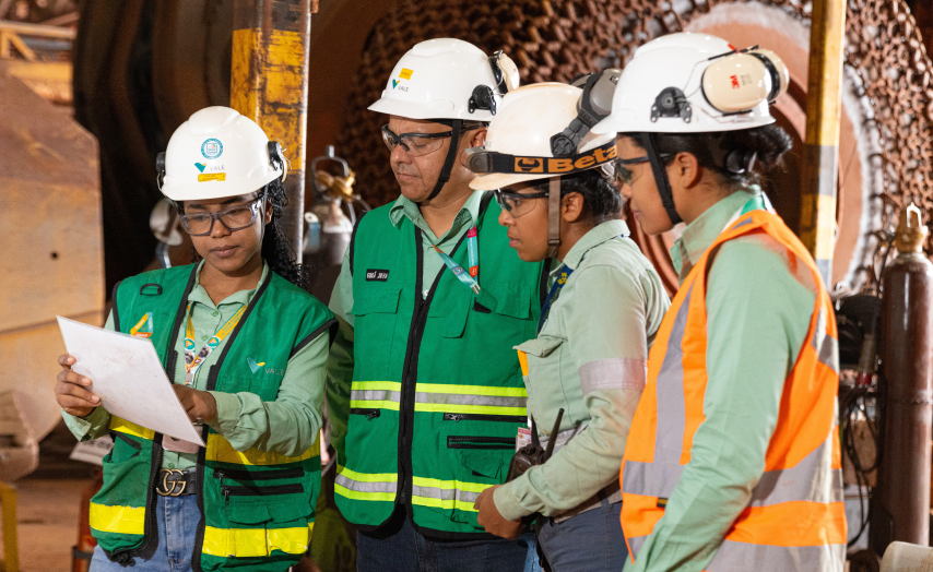Four Vale professionals wearing hard hats and safety vests review documents at one of the company's mining facilities.