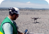 A man holding a control space and a drone in the background on a large, open, pebbled floor. The man is wearing a uniform, helmet and glasses.