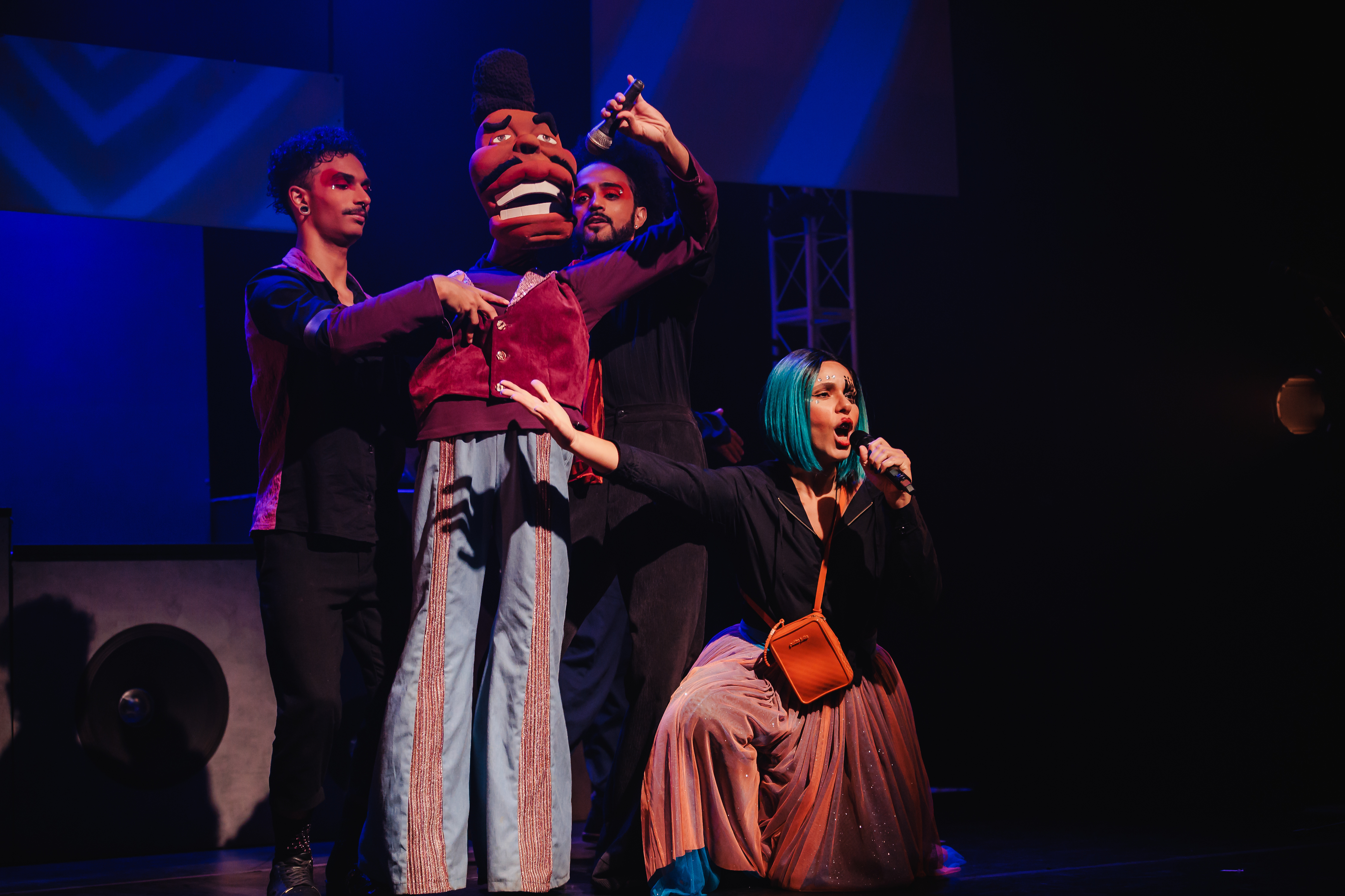 Dois rapazes seguram um boneco grande enquanto uma moça com peruca curta lisa está agachada na diagonal deles no palco, com o microfone em uma das mãos cantando.