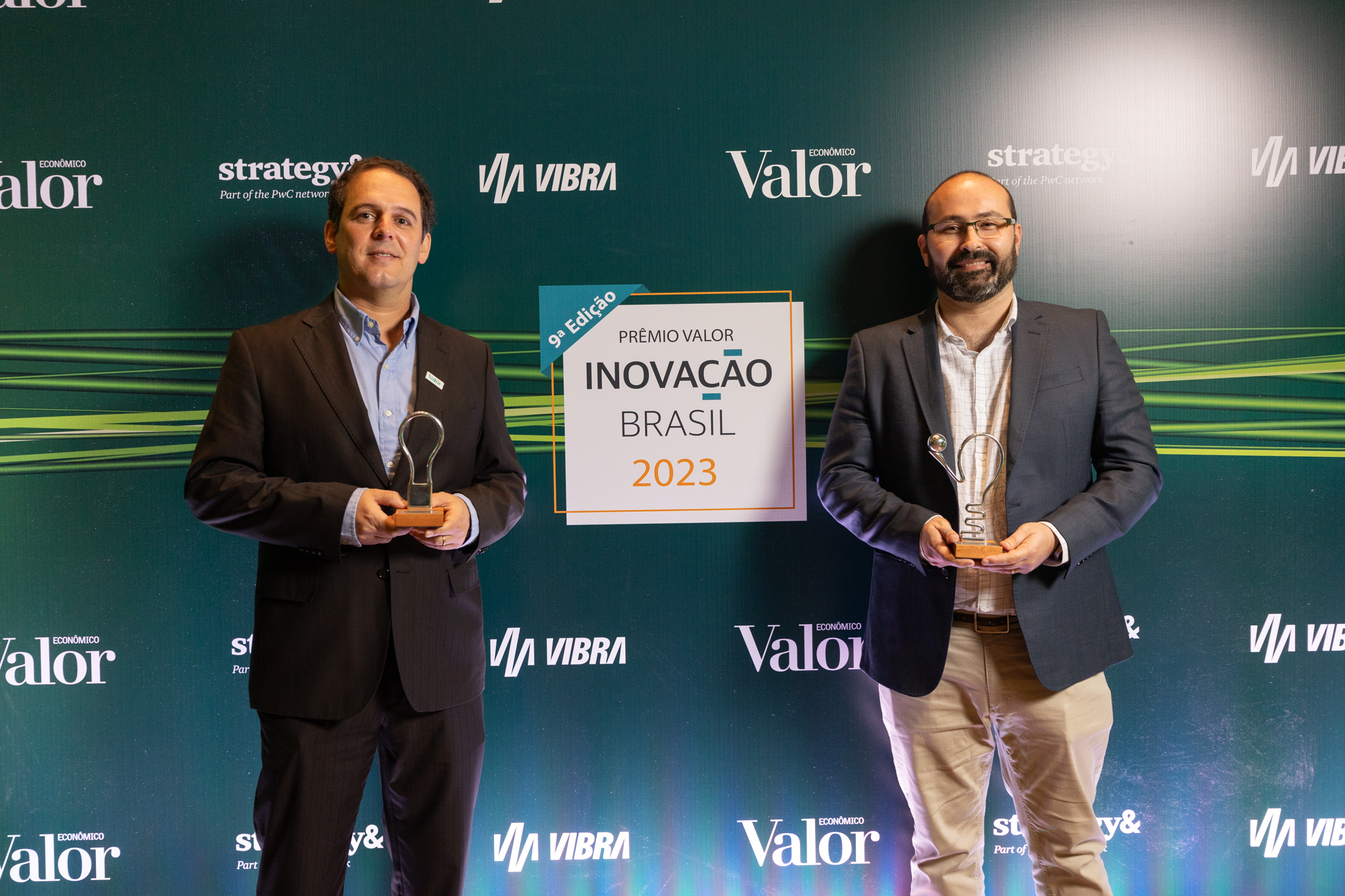 Dois homens usando roupas sociais seguram prêmios em frente a um painel onde está escrito: 9ª edição. Prêmio Valor Inovação Brasil 2023